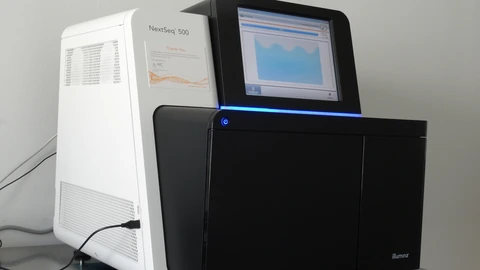 A white-and-black Illumina NextSeq 500 desktop sequencer on a bench, power cable at left, small screen showing a pale waveform, and the Illumina mark on the black door. No people appear.