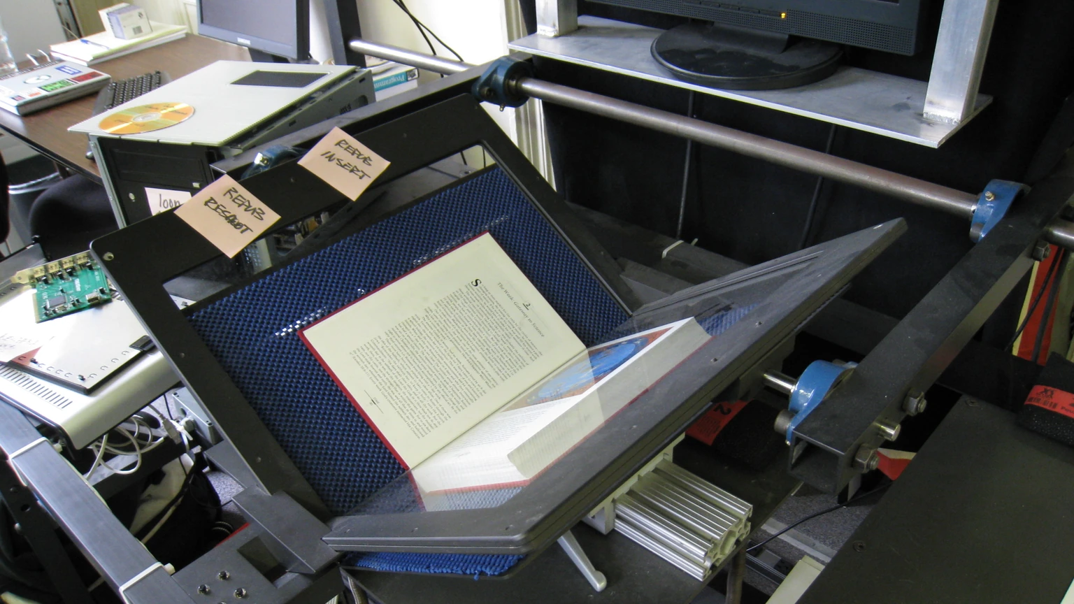 An open printed book on the glass platen of a black overhead book scanner, with two handwritten sticky notes on the frame and a monitor in the background.
