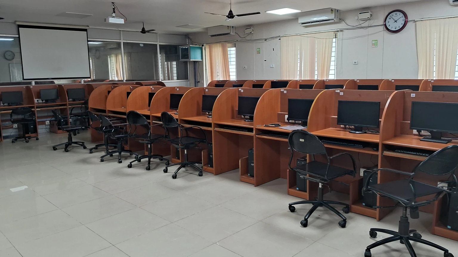 An empty computer laboratory with wooden desks, black chairs, dark monitors, a blank projector screen, and a white wall clock. No people appear.