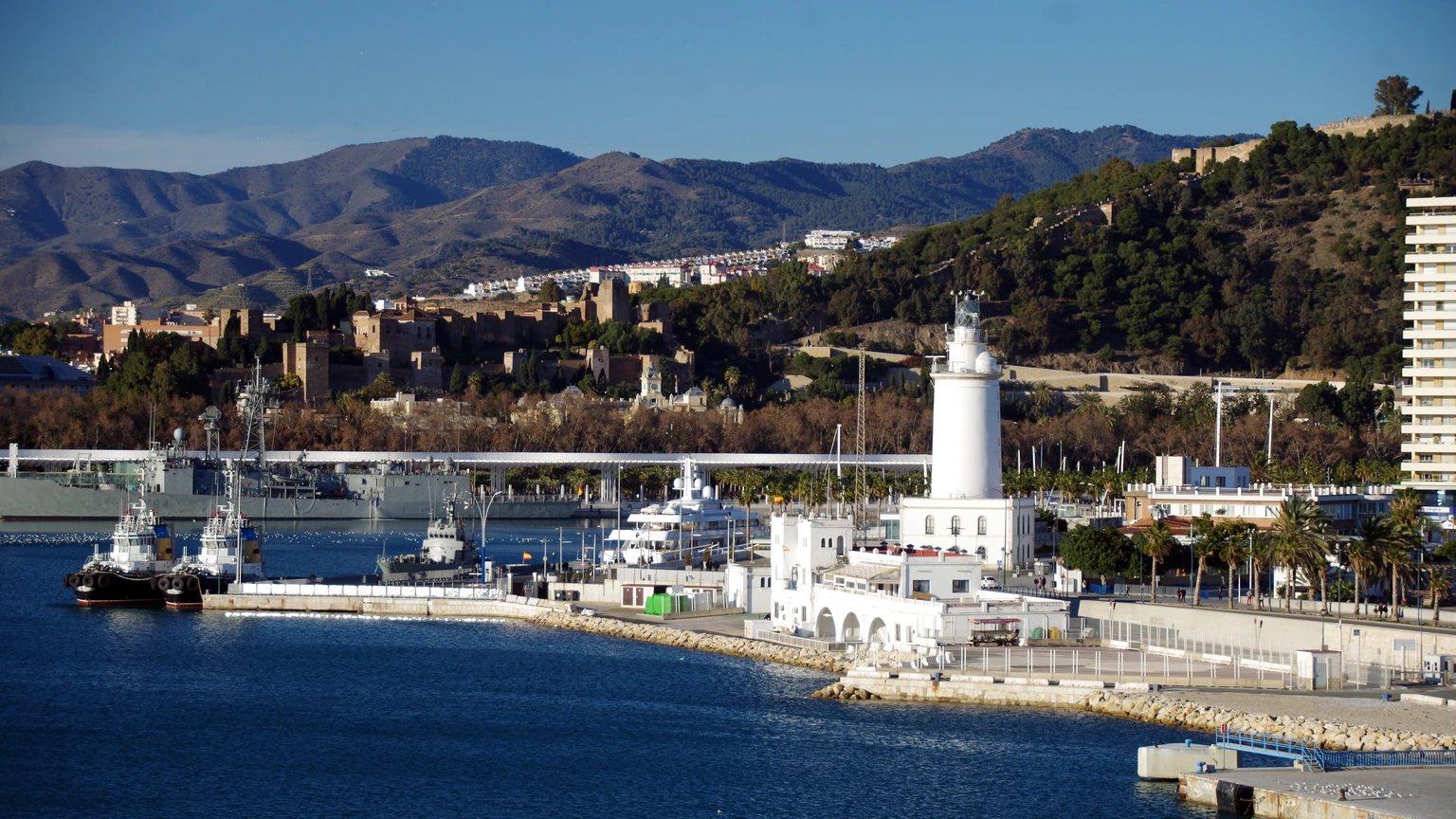 Málaga harbour on a clear day: deep blue water, a white lighthouse and harbour office, a black-hulled tug, and hills with scattered buildings. No identifiable people appear.