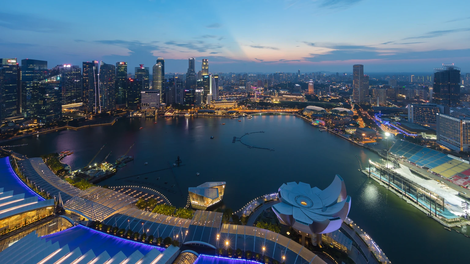 Aerial dusk view of Singapore’s Central Business District across Marina Bay, with the lotus-shaped ArtScience Museum and a curved bridge in the foreground. No identifiable people appear.