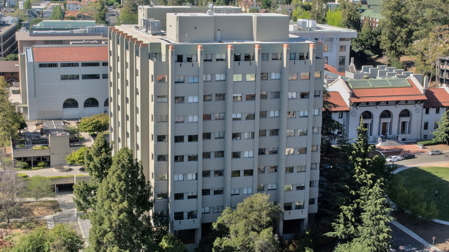 Aerial view of UC Berkeley’s beige high-rise Evans Hall from Sather Tower, trees at the base, a white classical building to the right. No people appear.