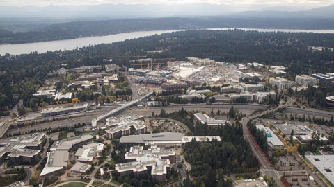 Aerial view of Microsoft’s Redmond campus under redevelopment, with office blocks, construction pads and a transit station at far left. No readable faces.