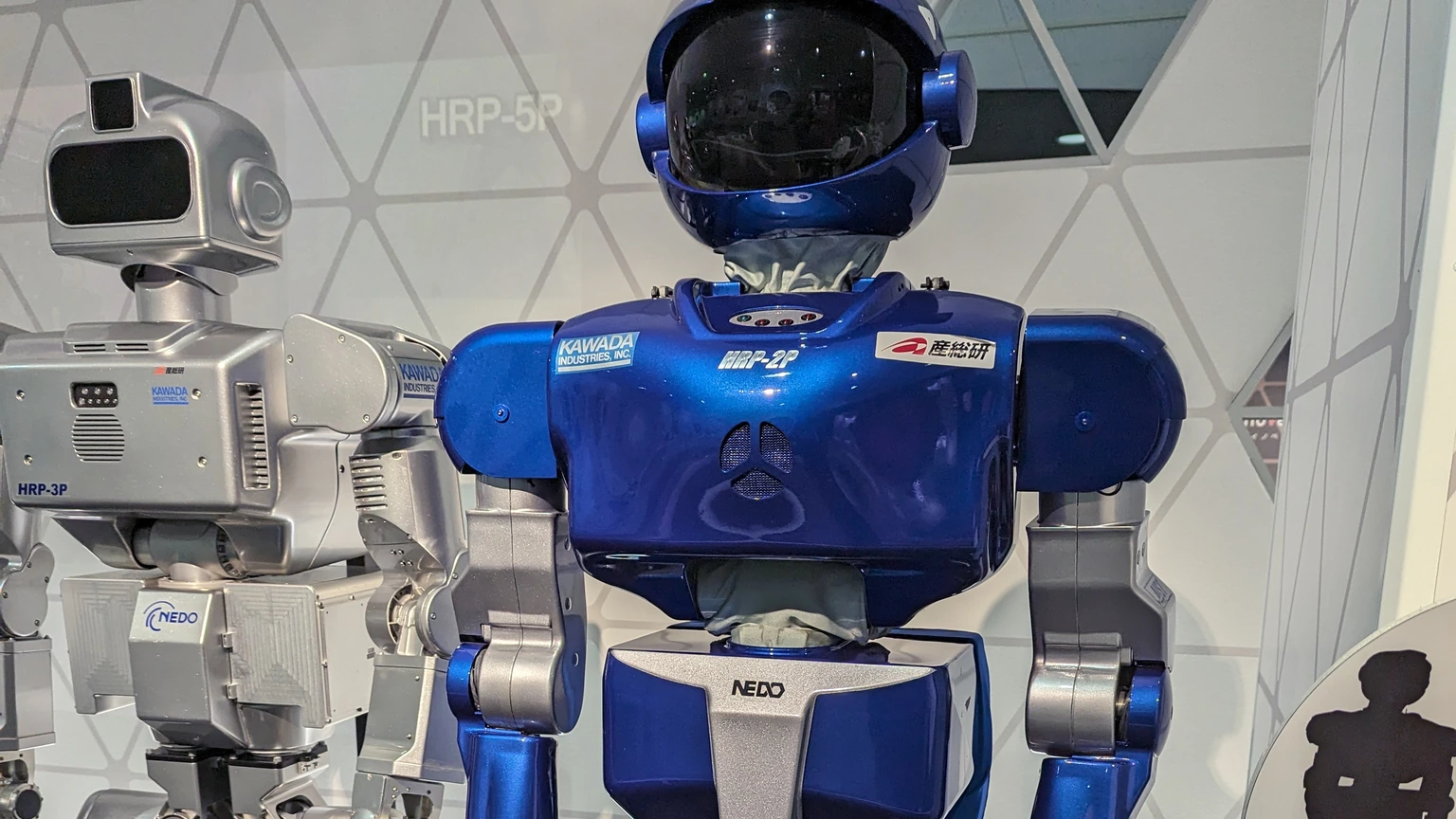 A blue Kawada HRP-2P humanoid robot standing next to a silver HRP-3P unit in a museum display, with an HRP-5P wall label behind them.