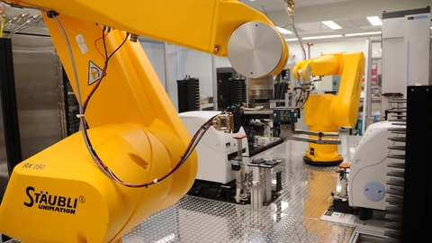 A yellow Stäubli Unimation RX160 robot arm in the foreground of a white lab, cable along the wrist, with a second yellow arm and white lab machines behind it. No people appear.