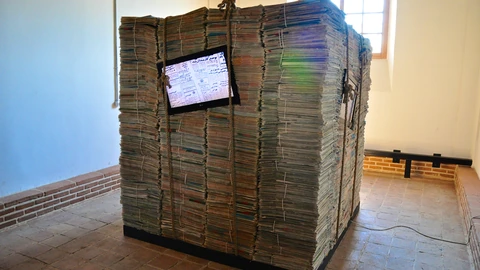 A cube of stacked newspapers tied with rope, with a small television set into one face, standing on a terracotta floor in a white room.