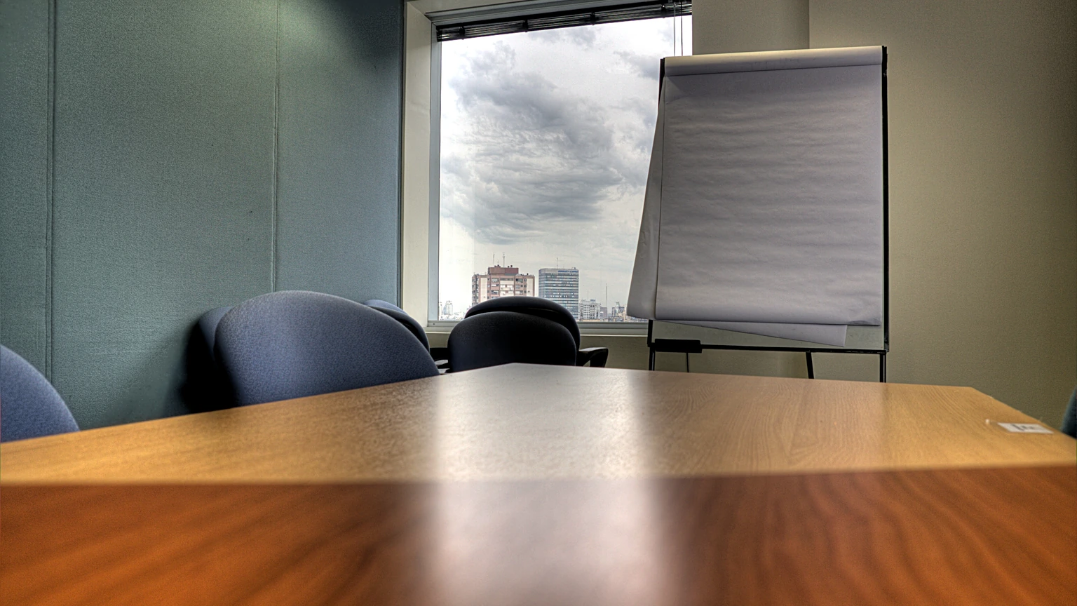 An empty meeting room: a pale wooden table in the foreground, two dark chair backs, a blank paper flip board, and a window with a cloudy sky. No people appear.