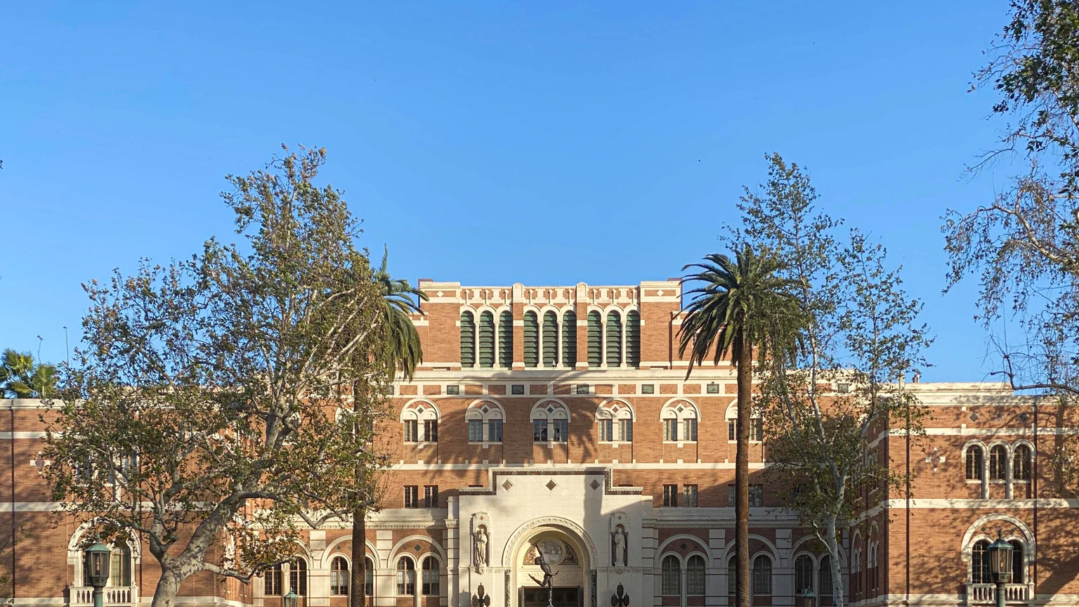 Upper brick facade of Doheny Memorial Library at USC under a clear sky, with palm trees in front of the entrance arch. No people appear in the crop.