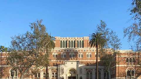 Upper brick facade of Doheny Memorial Library at USC under a clear sky, with palm trees in front of the entrance arch. No people appear in the crop.