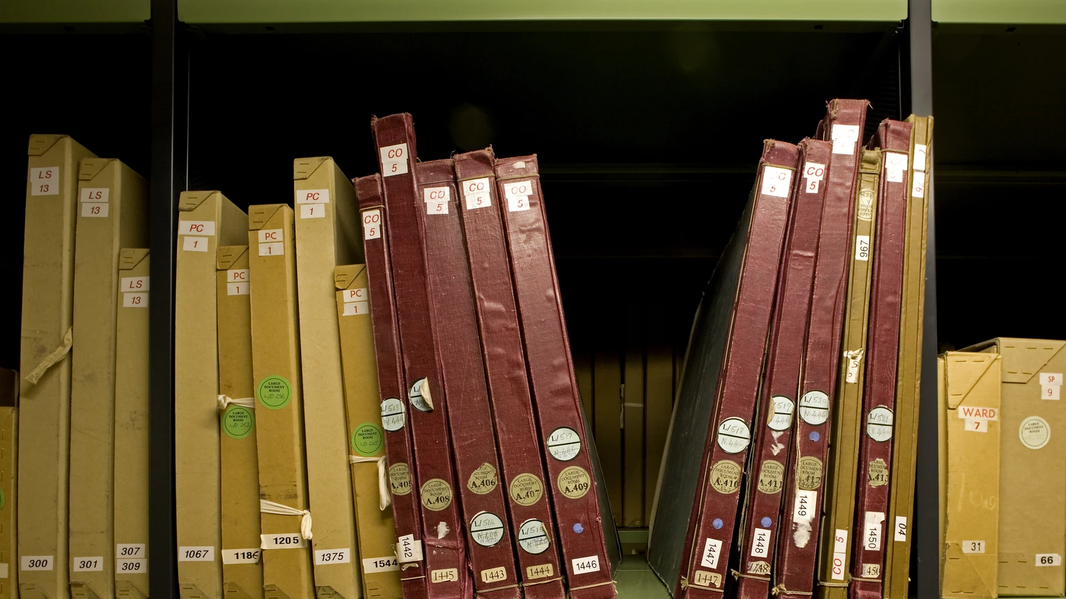 Cream archival boxes and maroon bound volumes labelled CO and PC on a metal shelf at The National Archives in Kew.
