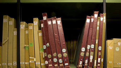 Cream archival boxes and maroon bound volumes labelled CO and PC on a metal shelf at The National Archives in Kew.