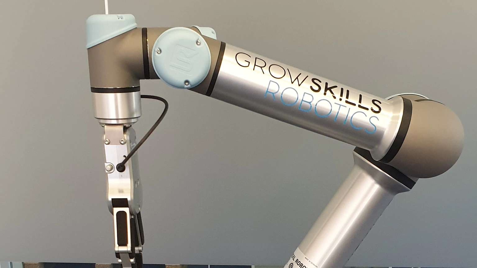 A silver-and-grey Universal Robots UR5 arm marked GROW SKILLS ROBOTICS, gripper down, against a plain grey wall. No people appear.