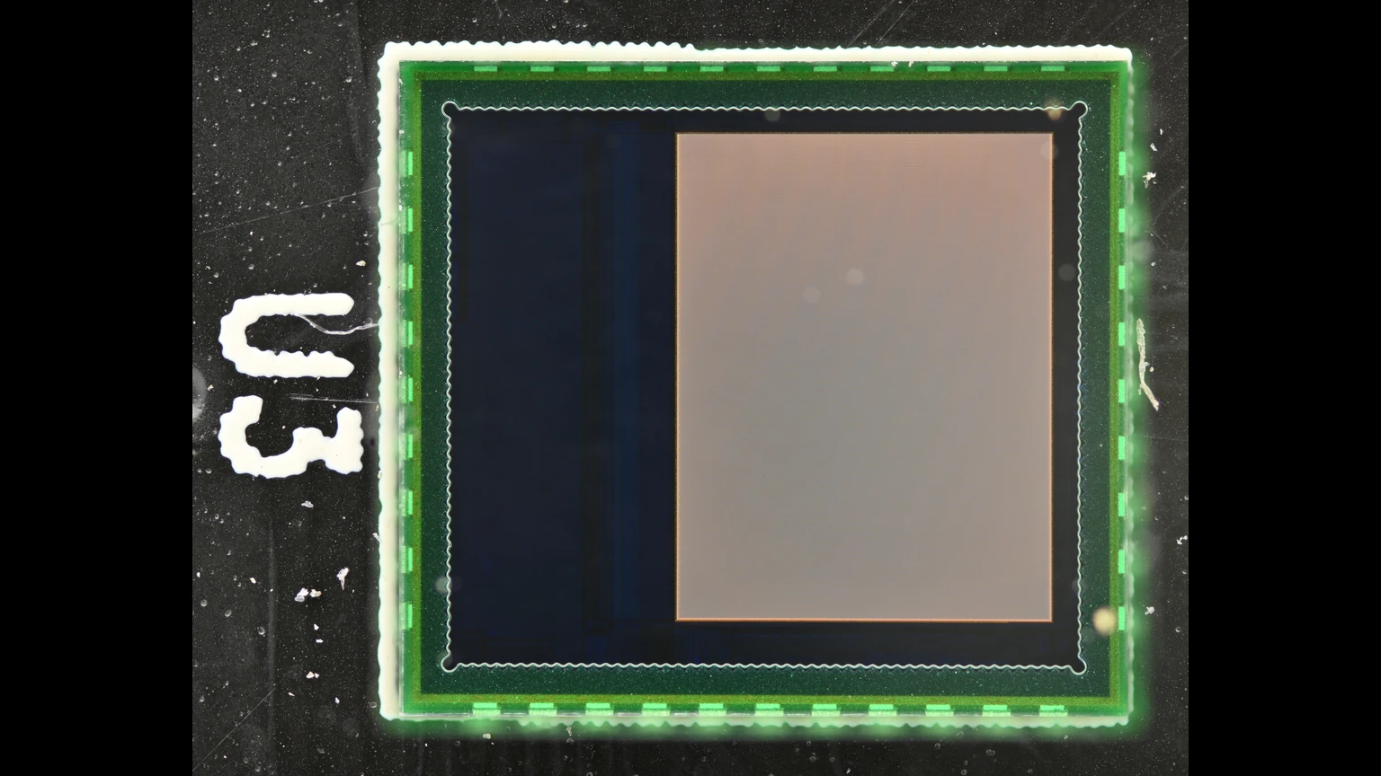Top view of a small CMOS image sensor in a glass chip-scale package on a black background, with a pale square active area, a green package rim, and a white U3 mark. No people appear.