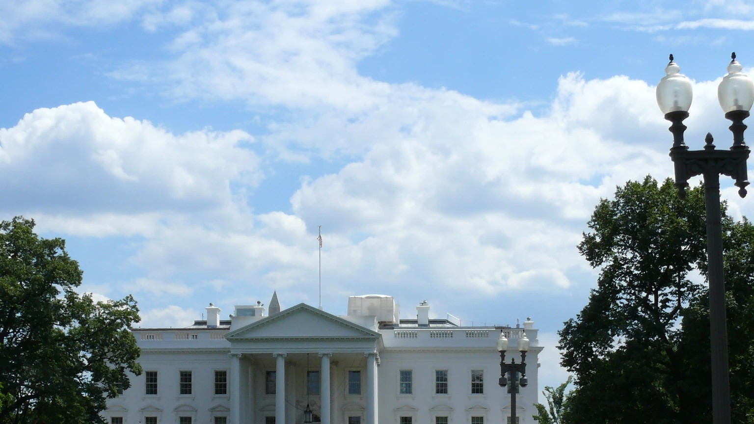 The White House north portico under a cloudy sky, with trees on both sides and a lamp post at right.