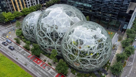 Aerial view of three glass geodesic domes beside an office tower and a wet city street with two distant cars. No identifiable people appear.