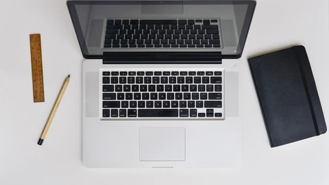 An open laptop on a wooden desk beside a keyboard, mouse, and notebook. No people appear.