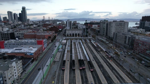 Aerial view of San Francisco’s 4th and King Caltrain station, with two silver trains on the center tracks, nearby streets, and the Bay Bridge beyond the skyline. No identifiable people appear.