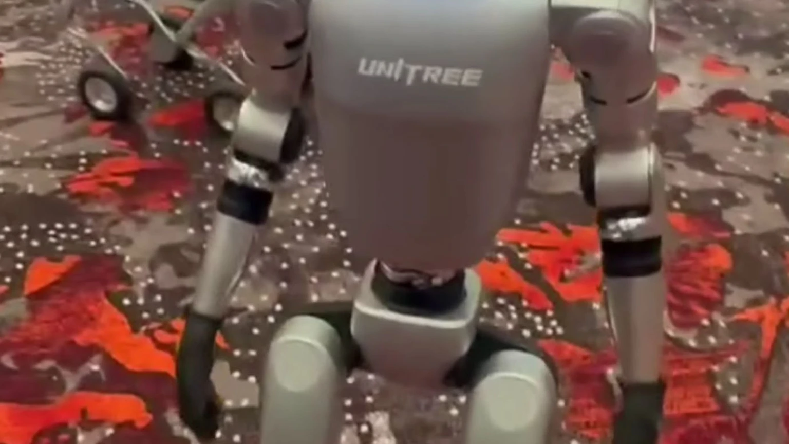 A silver Unitree G1 humanoid robot with a blue visor standing indoors. No faces are visible in the crop.