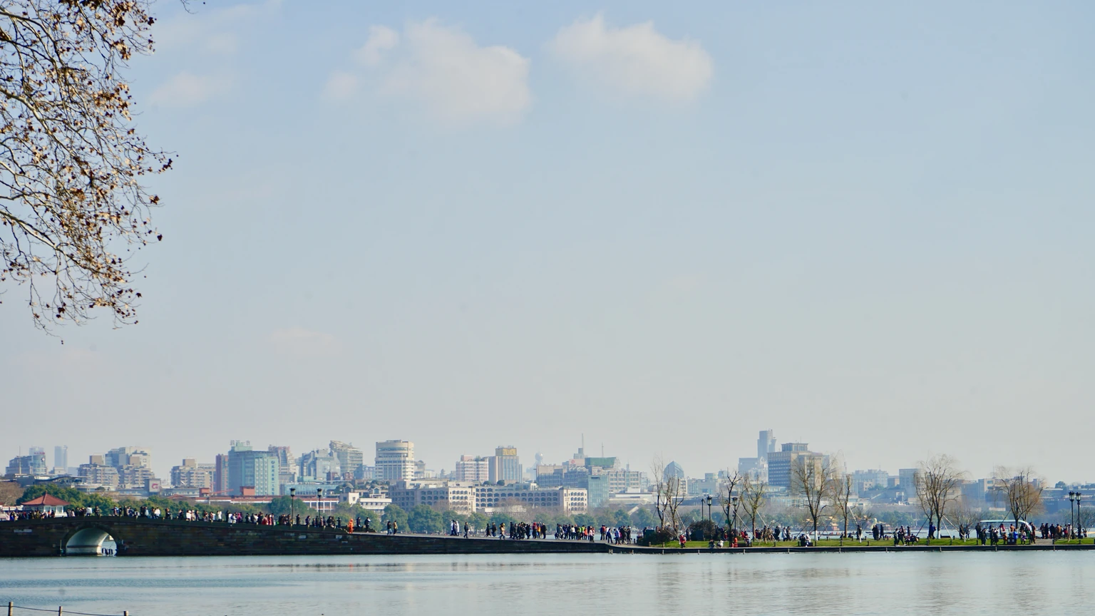 Hangzhou skyline of glass towers across West Lake under a clear sky. No identifiable people appear.
