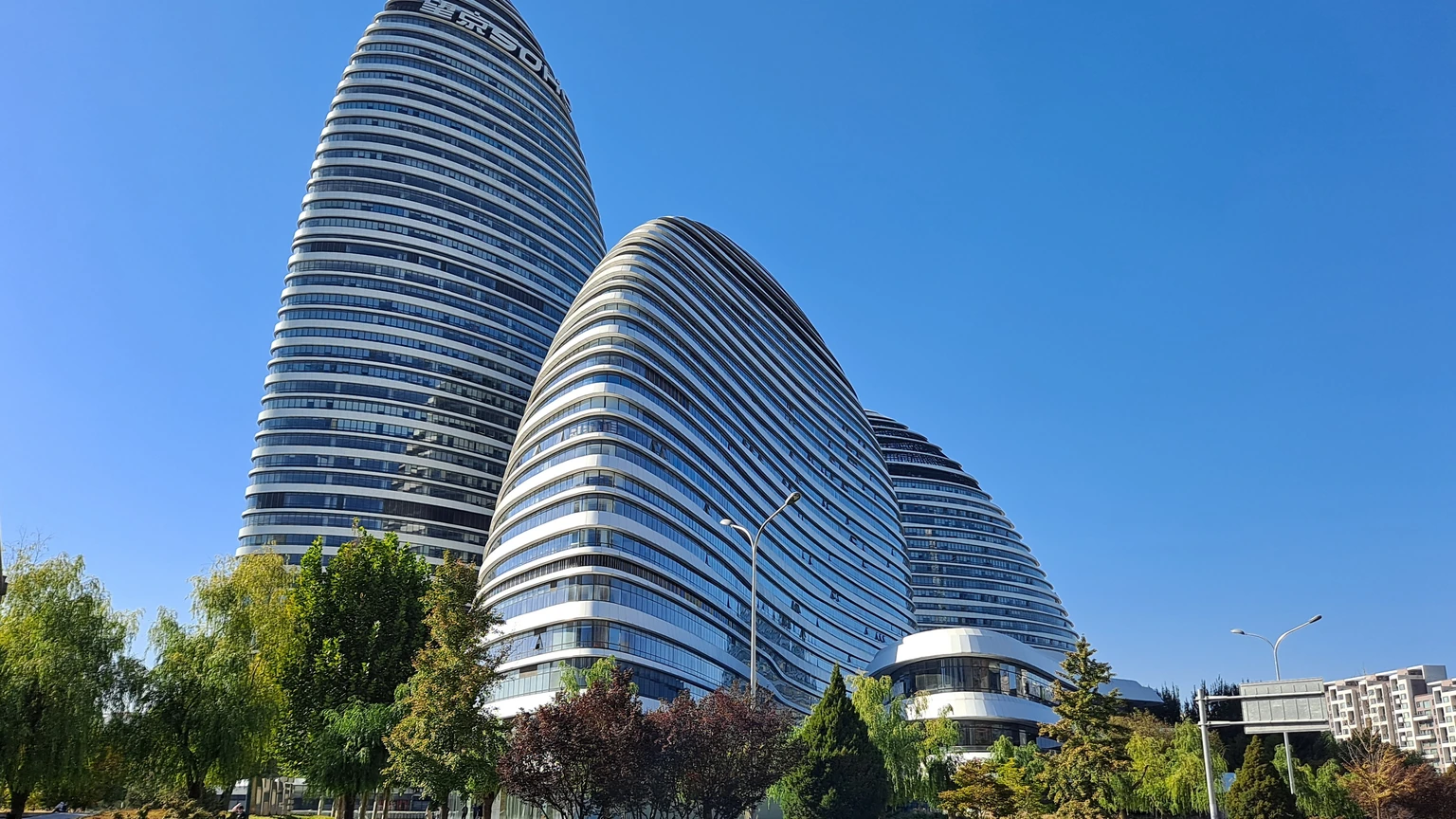Three white curved Wangjing SOHO towers against a clear blue sky, with street trees and a lamp post below. No people appear.