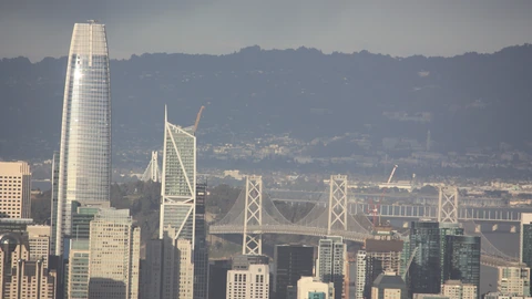 Hazy distant view of San Francisco’s skyline with Salesforce Tower, the diamond-braced 181 Fremont tower, and the Bay Bridge. No identifiable people appear.