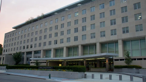 The pale stone south facade of the Harry S. Truman Building under a pale sky, with a curved canopy, a low HARRY S TRUMAN BUILDING sign, and an empty driveway. No people appear.