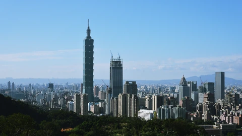 Taipei skyline with Taipei 101 rising above surrounding towers under a blue sky. No identifiable people appear.