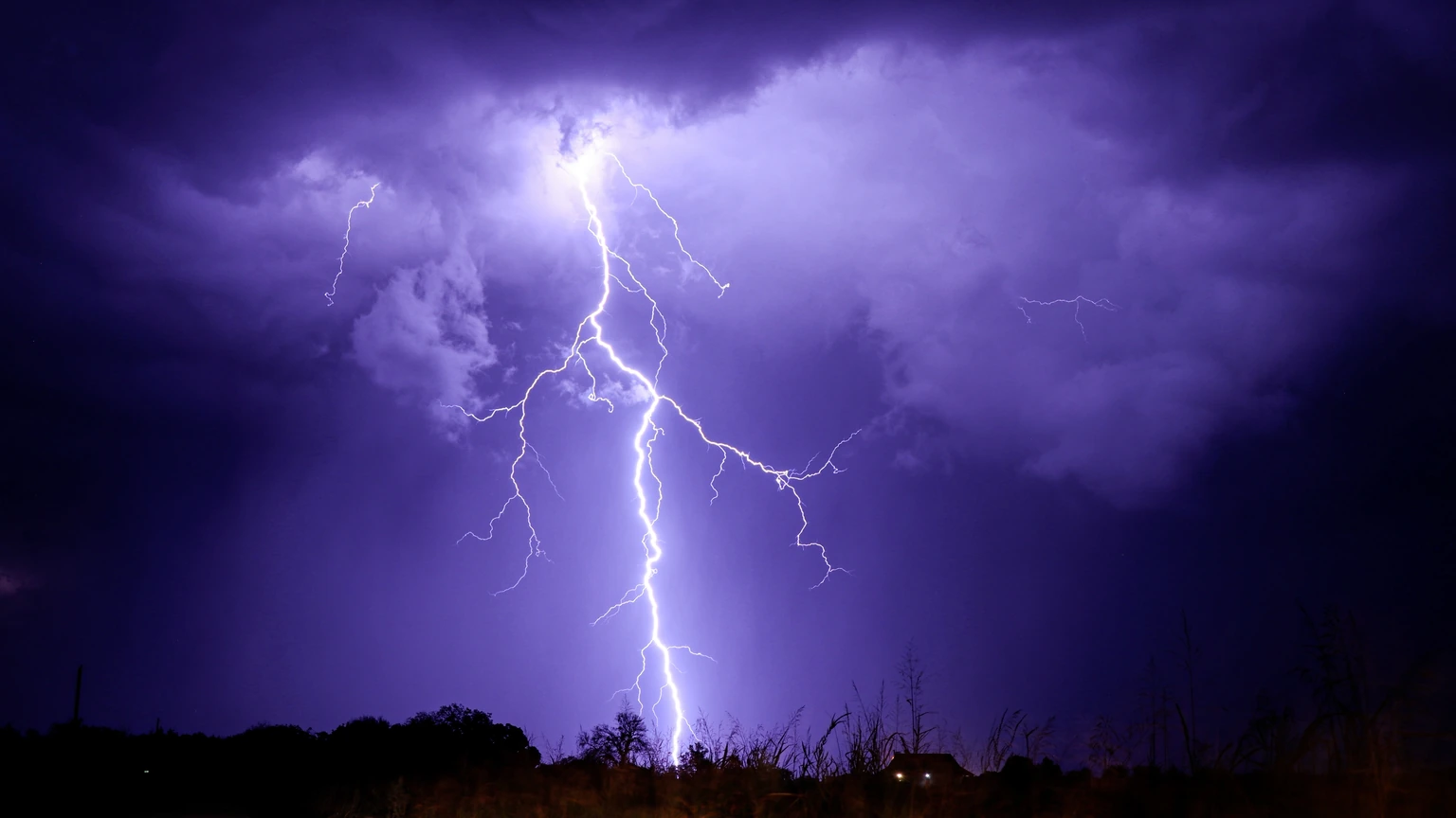 A bright cloud-to-ground lightning bolt over a dark landscape with illuminated clouds. No people appear.