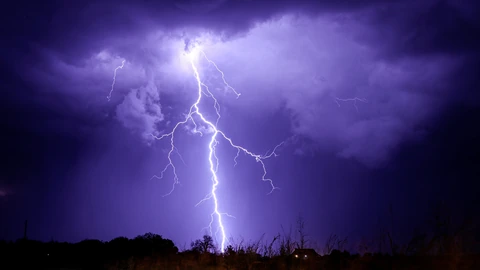 A bright cloud-to-ground lightning bolt over a dark landscape with illuminated clouds. No people appear.