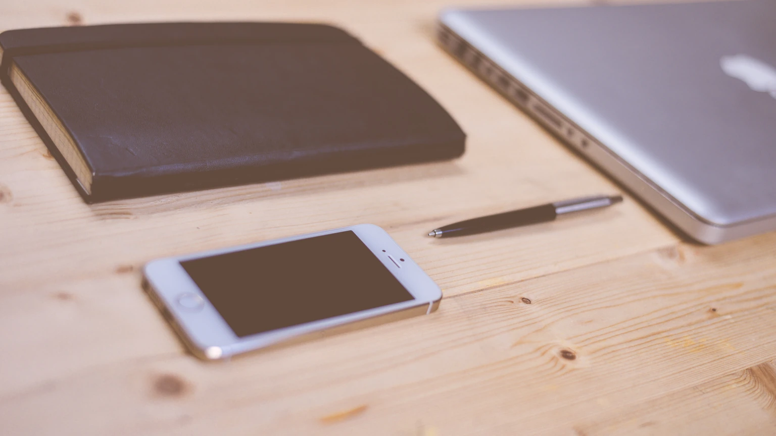 A white smartphone, pen, and notebook on a light wooden desk beside a laptop edge. No people appear.