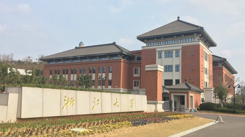 Stone wall sign reading Zhejiang University in gold Chinese characters, with red-brick campus buildings and dark tiled roofs behind flower beds. No people appear.