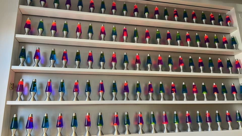 A tall white shelf wall filled with dozens of colorful lava lamps at Cloudflare's office. No people appear.