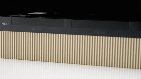 Close-up of the side of an NVIDIA H100 PCIe graphics card on a white surface, showing the H100 label, two NVLink connector covers and a gold ribbed heatsink.