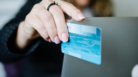 Close-up of a hand holding a blue payment card against the lid of a grey laptop during an online purchase; the background is softly blurred.