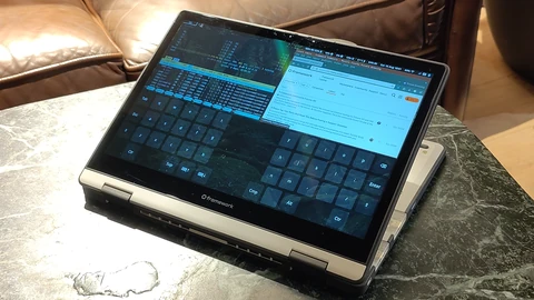 A convertible laptop folded into tablet mode on a marble table, its touchscreen showing a web browser, a terminal system monitor and a large on-screen keyboard.