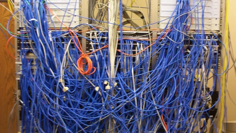 A server rack buried under a tangle of blue and orange network cables running between patch panels and switches.