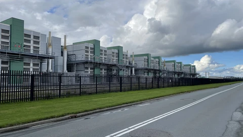Long row of grey and green Microsoft datacenter buildings behind a black steel fence, beside an empty road under a cloudy sky.