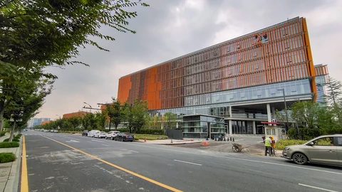 A large office building with an orange slatted facade and glass lower floors beside a wide road with parked cars under a grey sky in Hangzhou.