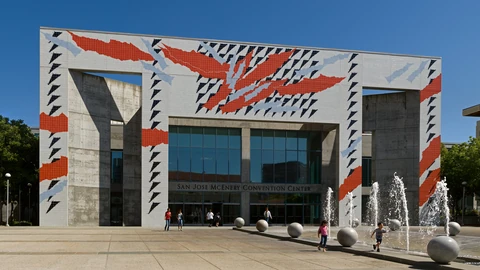 The concrete entrance of the San Jose McEnery Convention Center, decorated with a large red and black geometric mural, with fountains and stone spheres on the plaza in front under a blue sky.