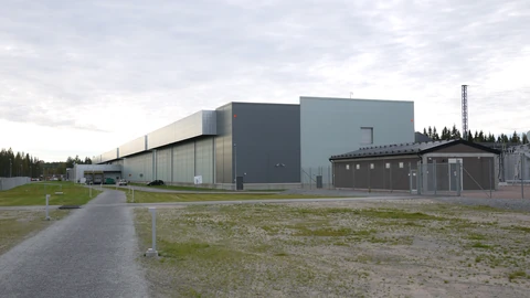 A long, windowless grey and pale-green data center hall beside a gravel path and grass field in Luleå, Sweden, with a smaller brown service building and fenced electrical equipment on the right under a cloudy sky.
