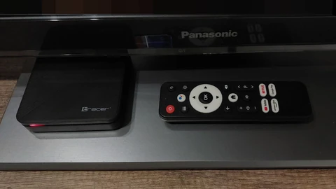 A small black Tracer Android TV set-top box with a red standby light, next to its remote control, on a glass TV stand under a Panasonic television. No people appear.