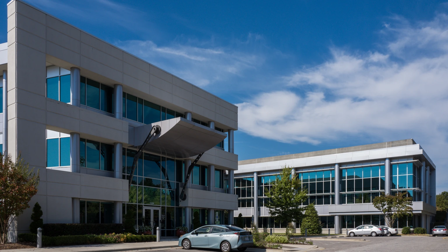 A three-storey office building with pale grey panels and blue-tinted windows under a blue sky with white clouds, a second glass-fronted wing behind it, trees and a parked car in the foreground.