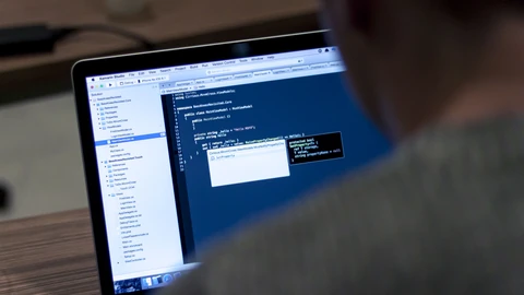 Over the shoulder of a person in a grey jumper, a laptop screen shows an IDE with a project file tree on the left and C# code in a dark editor pane, with an autocomplete pop-up open.