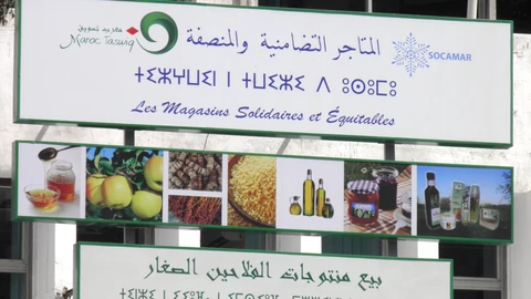 Two white shop signs with green borders on a building in Morocco. The upper sign repeats the same store name in Arabic script, in Tifinagh script and in French, next to a green swirl logo and a blue snowflake logo; below it a strip of photos shows honey, apples, couscous, olive oil and jam.