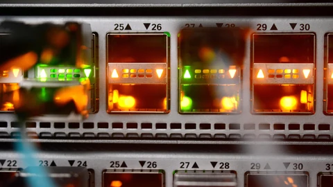 Close-up of a network switch in a computing centre: rows of numbered ports, 25 to 30, glowing orange and green, with a blurred cable plugged into one port in the foreground.