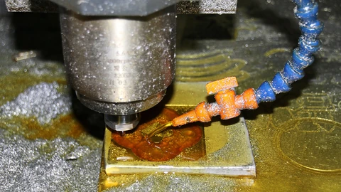 Close-up of a CNC milling machine: a steel spindle with a small cutter lowers onto a brass plate, cutting a rounded shape, while an orange coolant nozzle on a blue segmented hose sprays fluid onto the cut and metal chips cover the bed.