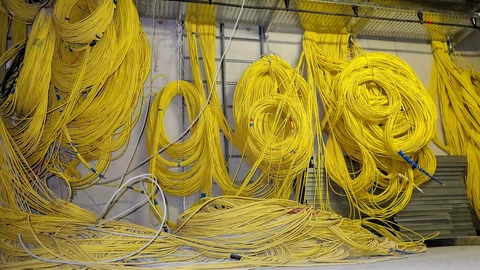 Dozens of coils of yellow fibre-optic cable hang in loops from racks along a server-room wall, with grey and blue cables running beneath them.