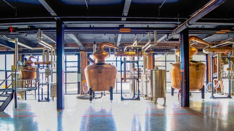 Three copper pot stills of different sizes with curved swan-neck tops stand on a polished distillery floor beneath a ceiling of pipes, alongside steel tanks.