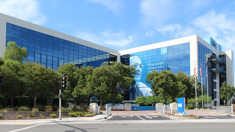 Intel’s blue glass headquarters building in Santa Clara, California, behind a row of trees, with a large banner reading ‘What can be done, can be outdone’ on its facade and an Intel sign at the entrance.