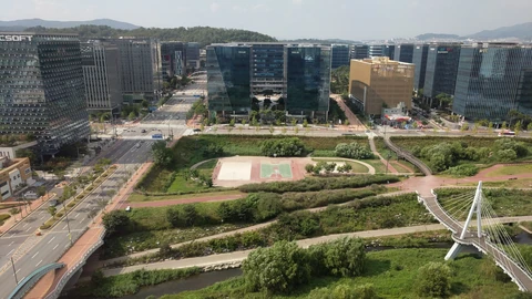 Aerial view of Pangyo Techno Valley in South Korea: glass office towers line a road behind a green park, sports courts and a white cable-stayed footbridge over a stream, with hills behind.